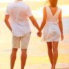 Young couple holding hands at beach sunset enjoying romance and sun. Young happy couple in love on romantic summer holidays vacation. Young lovers in casual clothing. Asian woman, Caucasian man.